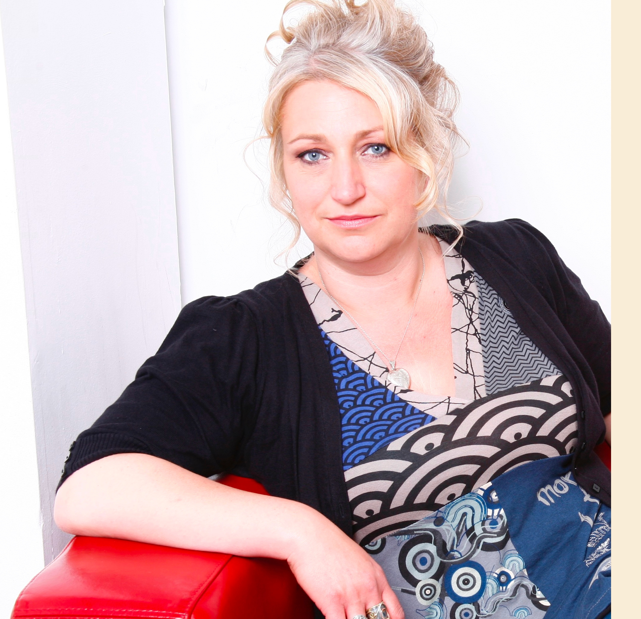 A photo of the artist, Zoe Partington. She is a white woman with blonde hair in an up do, wearing a blue dress and black shawl. She is sat on a red sofa against a white wall, looking into the camera.